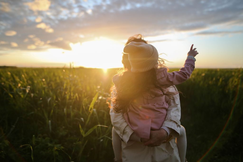 A woman gives her young daughter a piggyback ride through a field at sunset.
