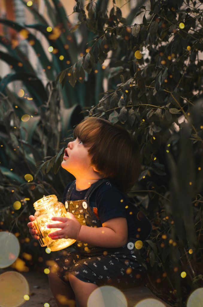 A little boy holds an open jar of lightning bugs and stares with wonder as they fly around him.