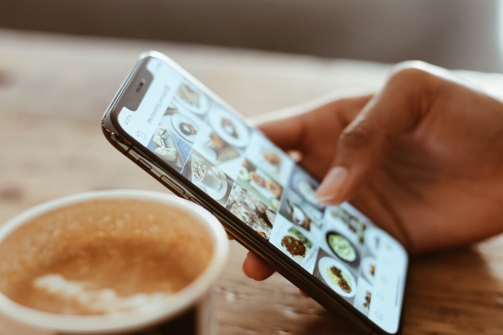 A woman's hand holding her phone, as she sips her nearby latte, posting about her favorite local coffee shop on Instagram.