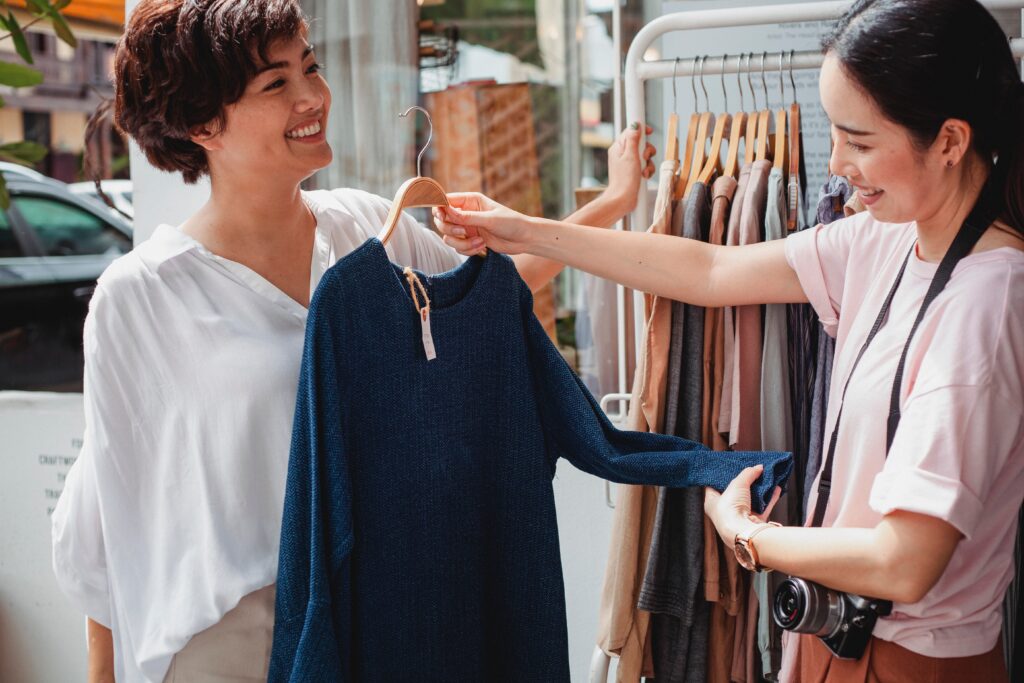 A smiling local shop owner helps a woman customer find a top in a friendly exchange.