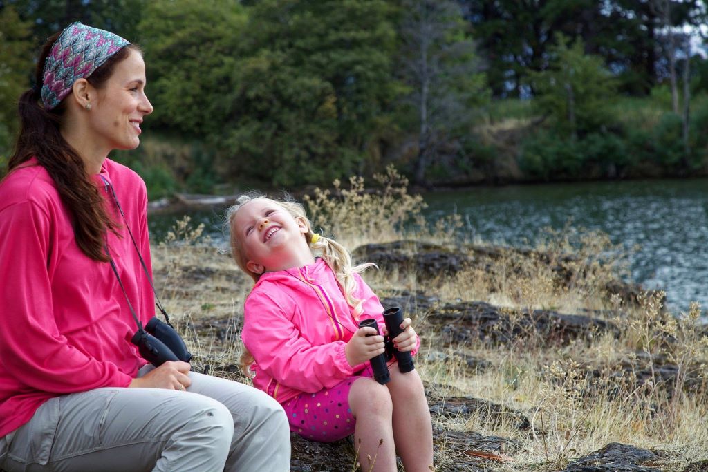 A woman and her young daughter sitting by a lake, birdwatching with binoculars, and smiling at one another.