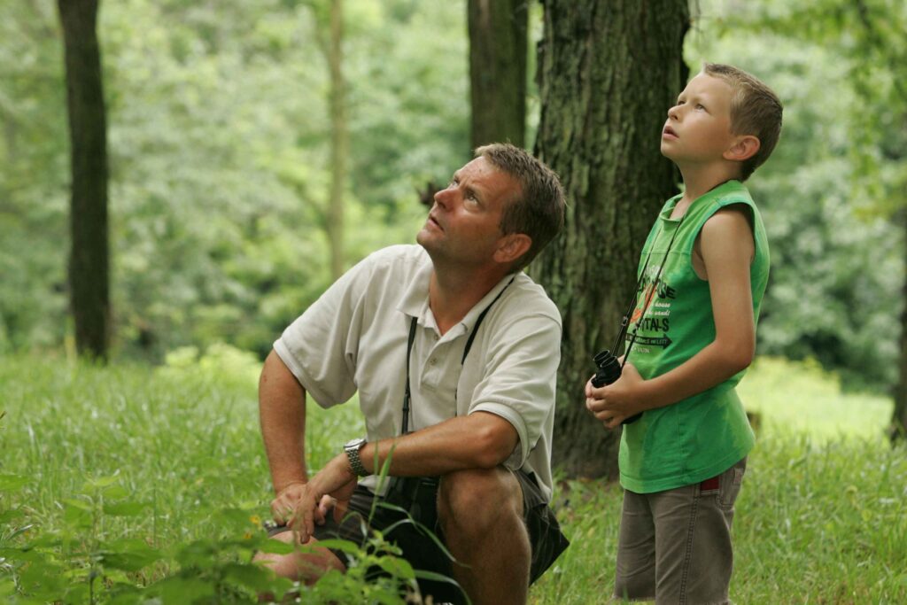 A father and son birdwatching in a forest, looking upward at a bird with their binoculars in their hands.