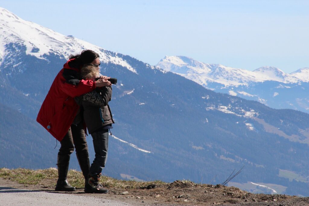 A woman adjusts her daughter's binoculars, with snow-capped mountains as their background. 