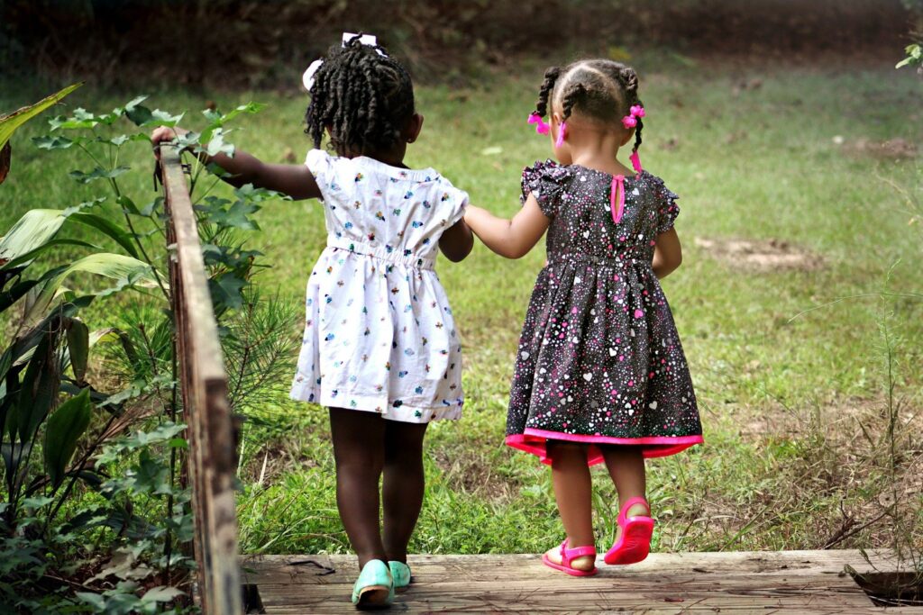 Two young girls hold hands as they walk away from the camera, into a green meadow.