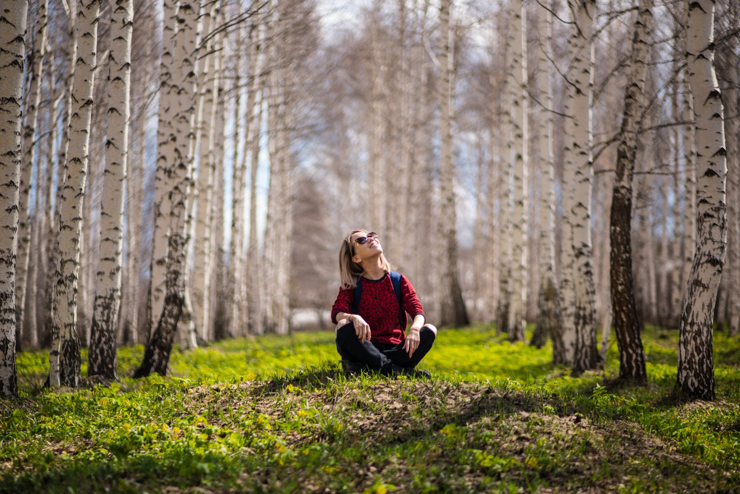 A smiling woman sits in a forest of aspen trees.