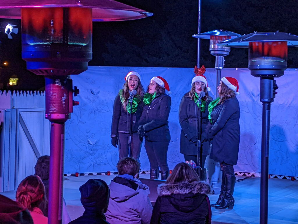 4 women singing on a stage wearing santa hats