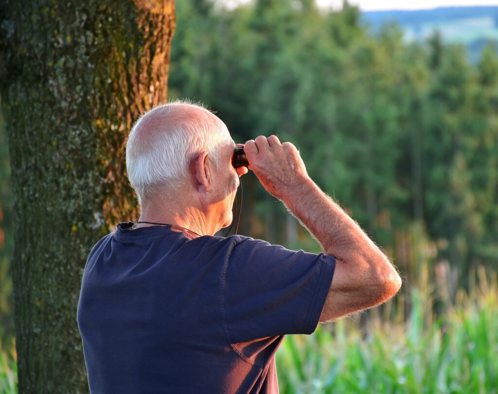 A white-haired man peers through a pair of binoculars at a bird.