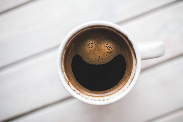 A cup of coffee with the bubbles in the shape of a smiley face, on a white wooden table shown from above.