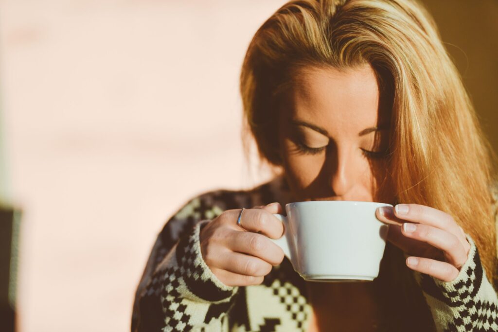 A blonde woman sips and savors a cup of coffee out of a white ceramic mug in a sunlit room.