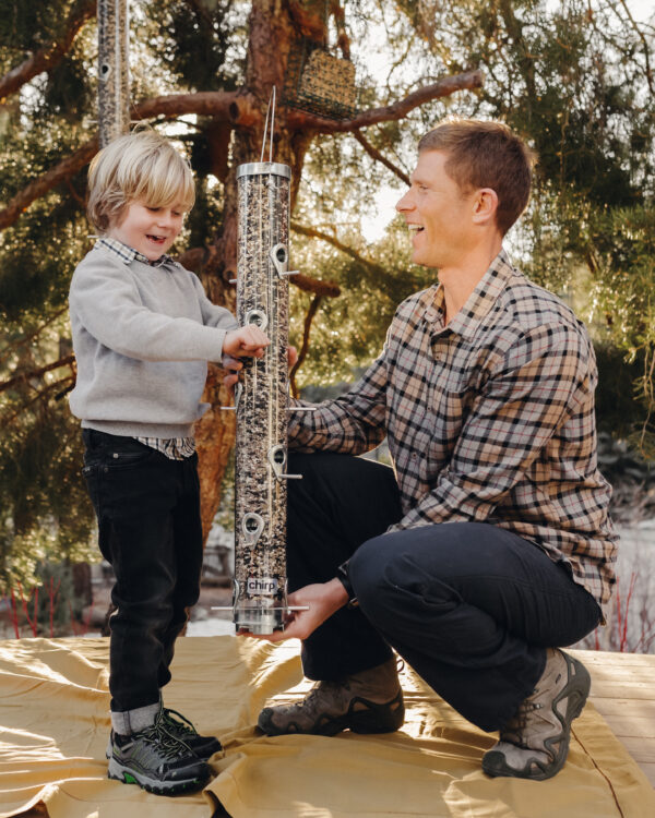 A man and his young son smile as they hold a large tube feeder full of Chirp's Big Bear Blend seed mix. 