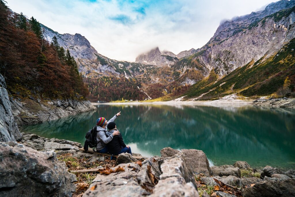 A couple sits on rocks near a beautiful lake, as the man smiles and points to something in the distance.