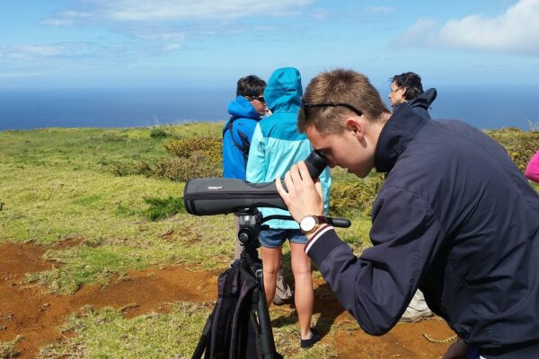 A young man looks through a birding optic in a natural landscape, with a bunch of birders, ocean, and greenery in the background.