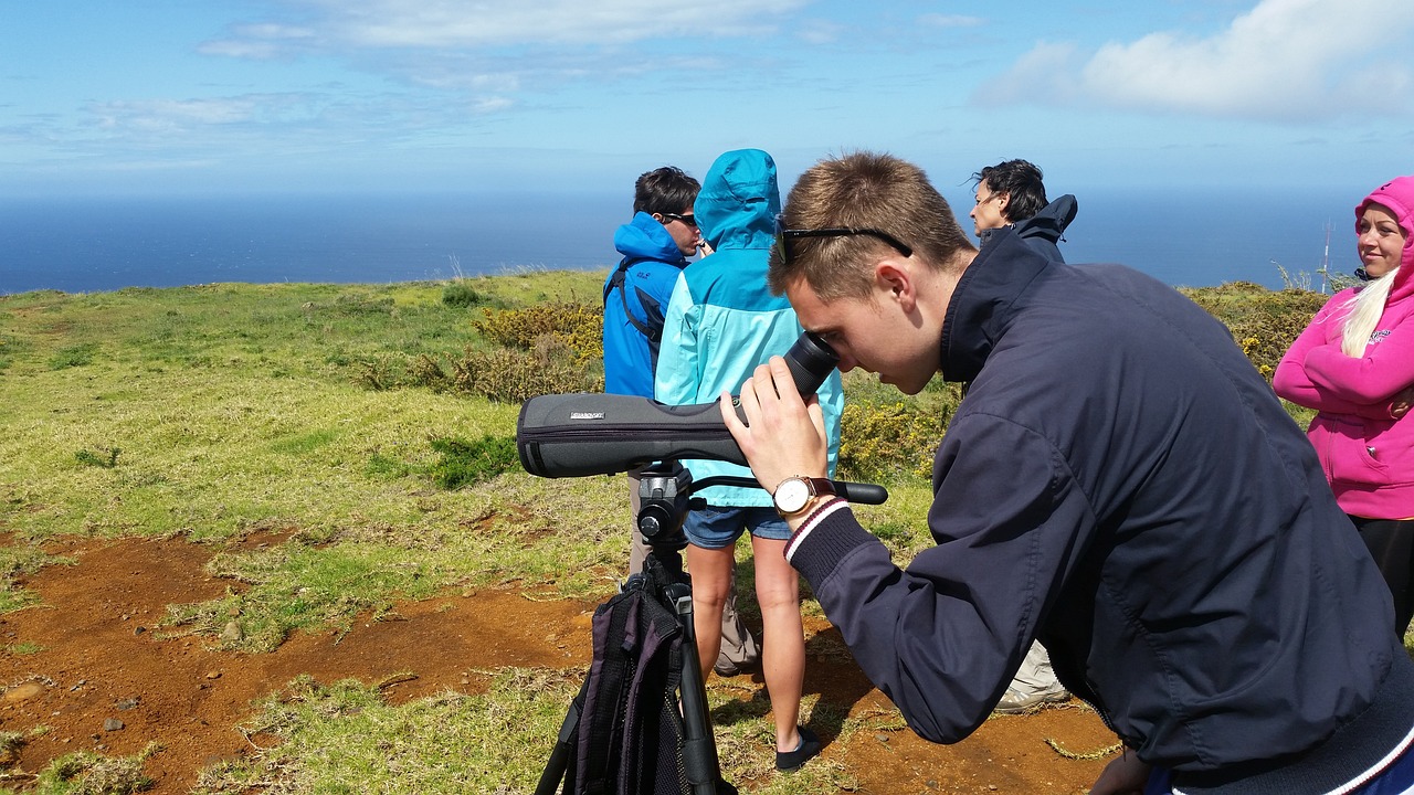 A young man looks through a birding optic in a natural landscape, with a bunch of birders, ocean, and greenery in the background.