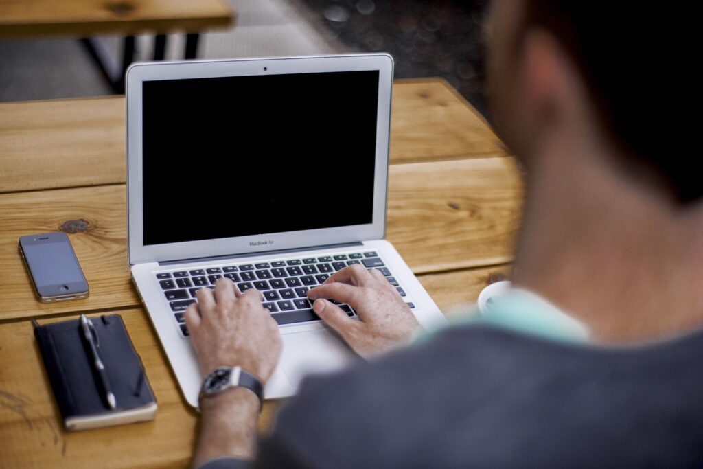 A man sits outside at a wooden table, working on a tablet with a phone and notepad next to him.