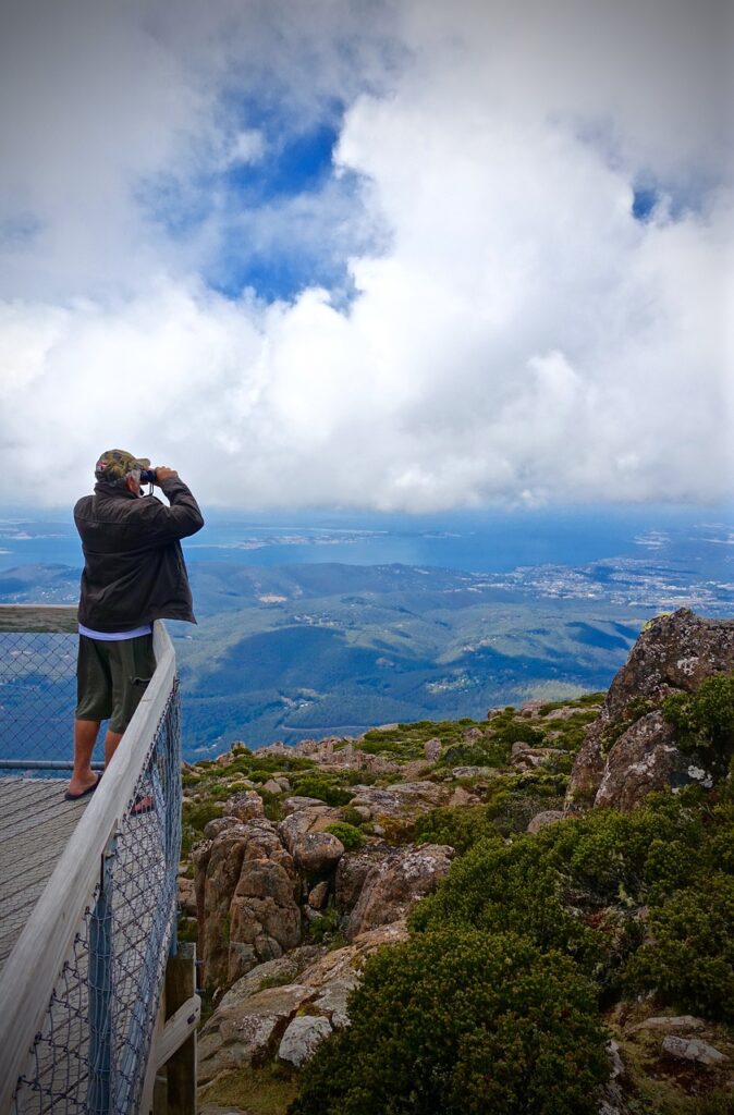 A man on an outlook point looks out over a mountainous landscape with a pair of binoculars.