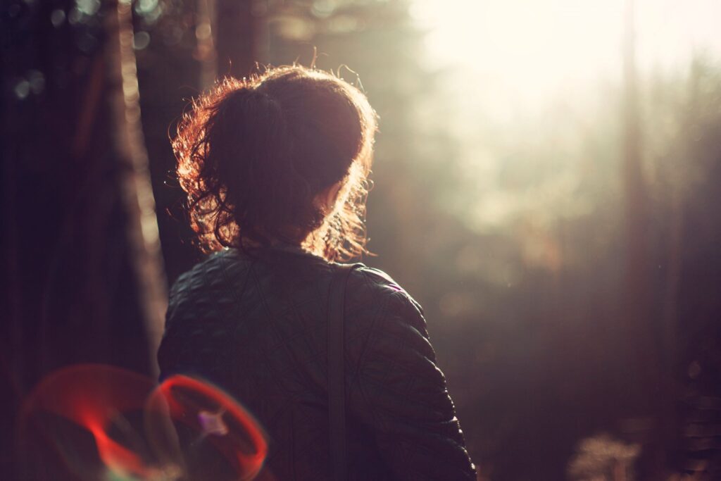 A woman shown from behind, sitting out in nature as the sun lights up the background.