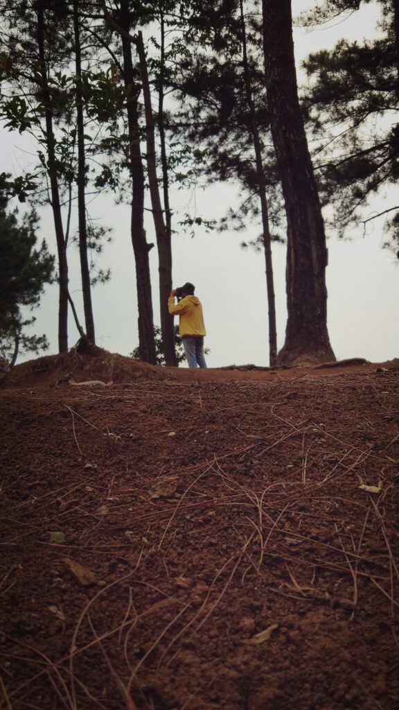 A man with a yellow jacket and hat stands in a wooded area, peering through a set of binoculars at a faraway spot. 