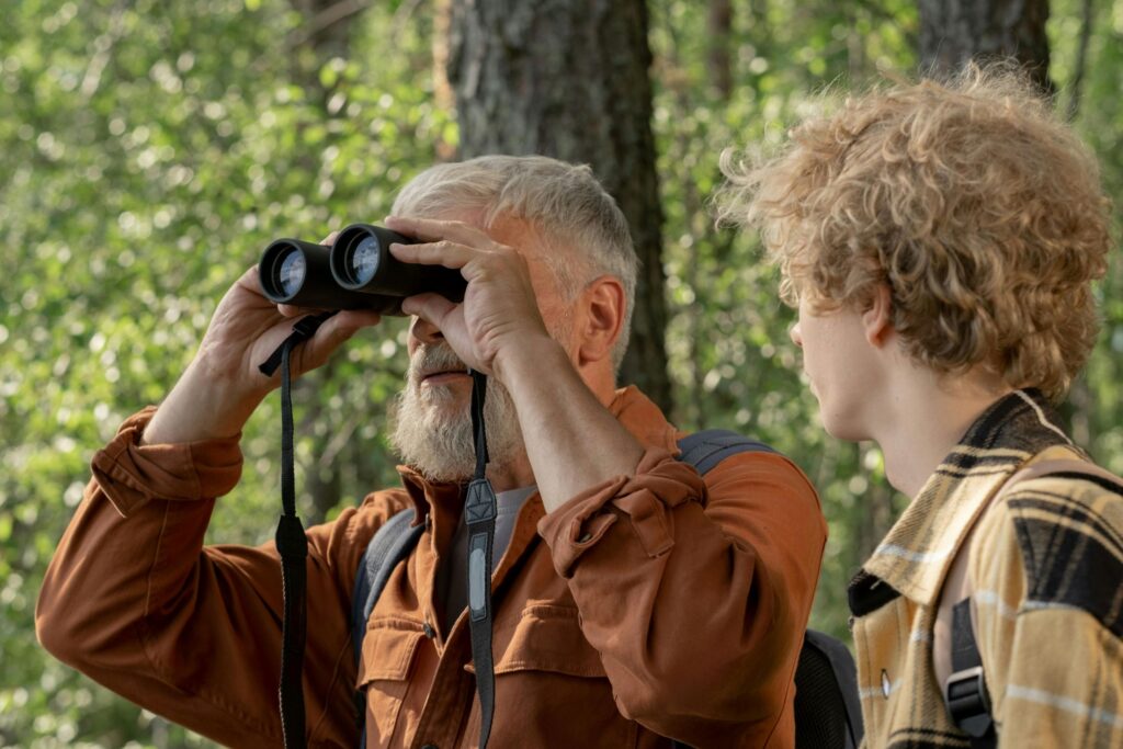 An older, white-haired man looks up through binoculars in a natural outdoor setting; a teenage boy looks at the man expectantly.