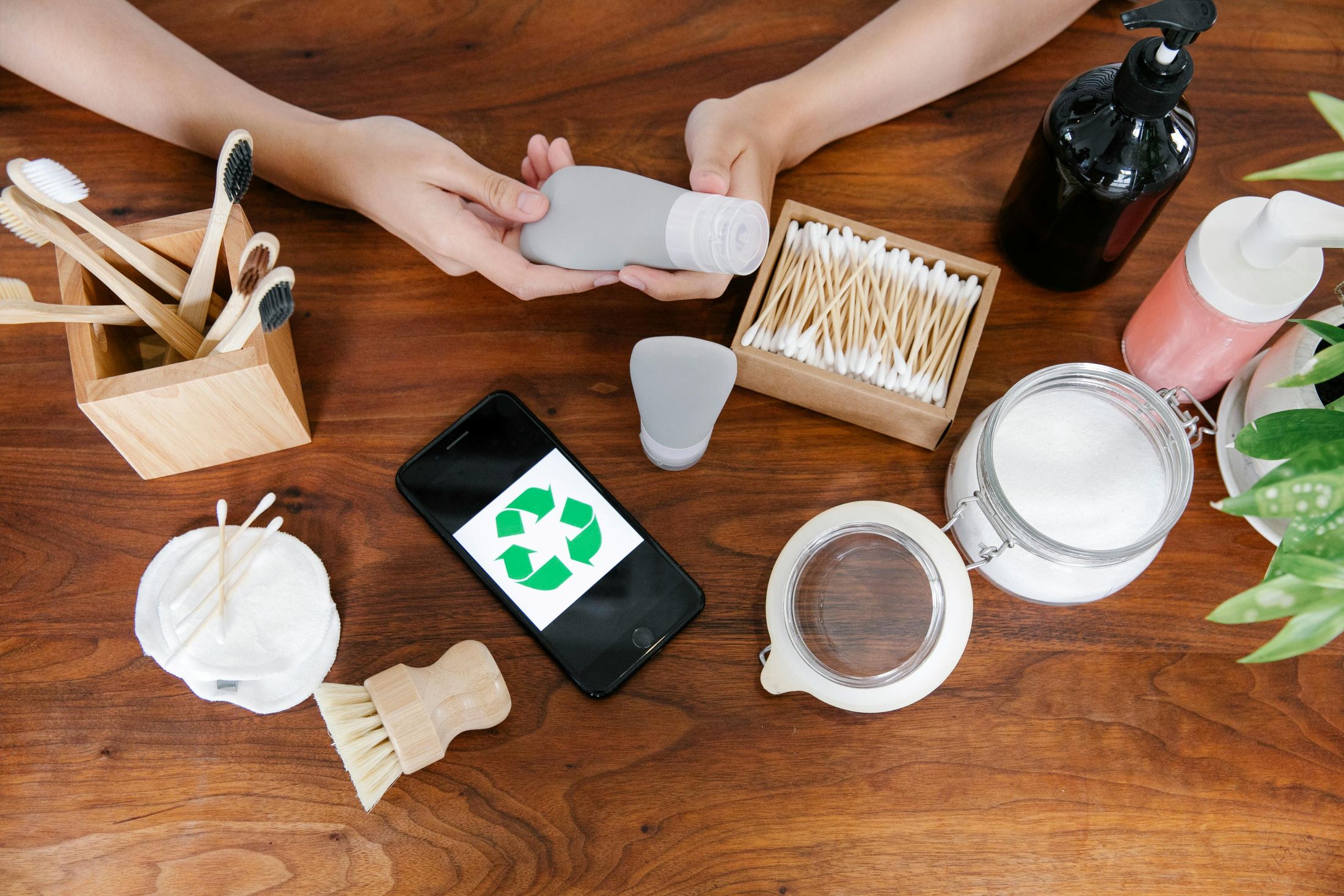 A wooden table is shown from above, with several items on it. A person's hands are holding a reusable plastic container, and there are bamboo and glass containers on the table.