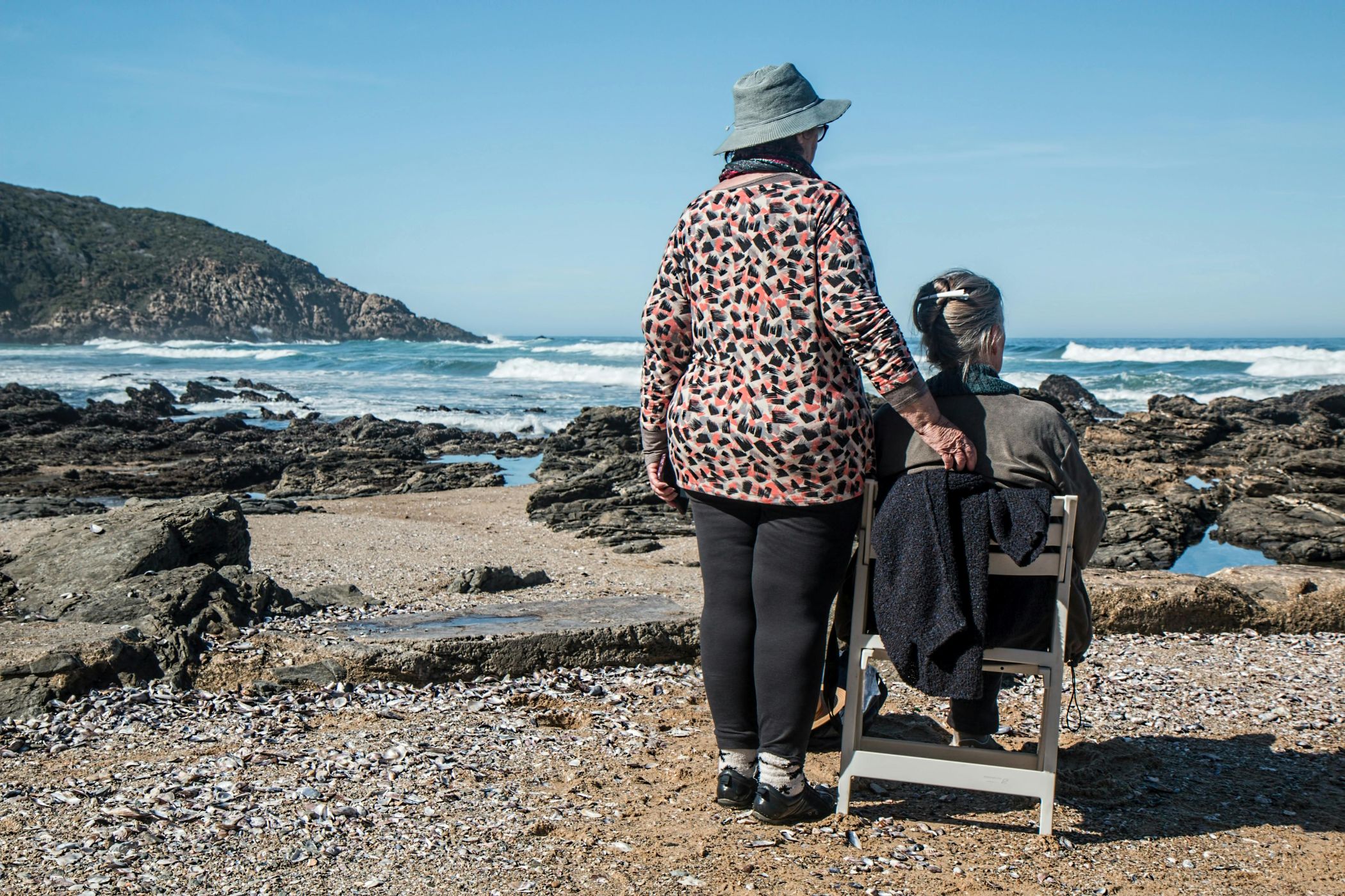 Two senior women, one sitting in a chair and one standing next to it. look out across a beach landscape.
