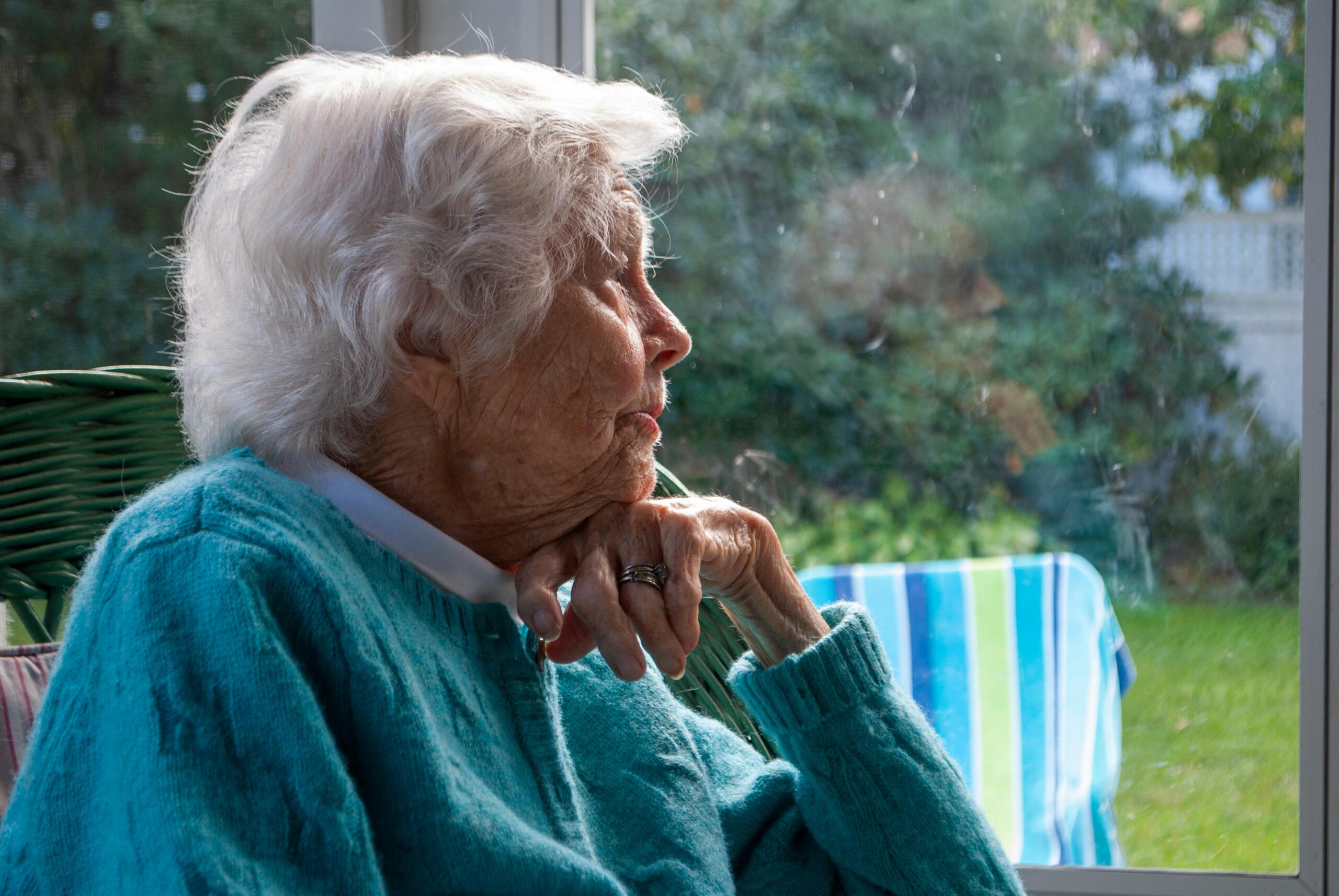 An older woman with white hair and blue sweater smiles as she looks out her window to her yard outside, presumably looking out at her busy bird feeder.
