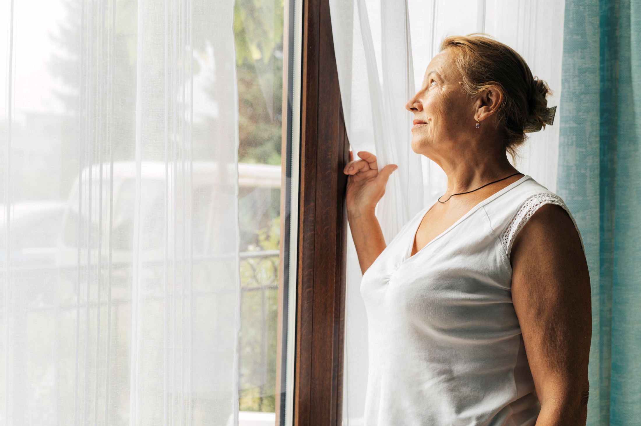 An older woman looks out her window and smiles as she observes the bird feeder hanging outside, and out of view.