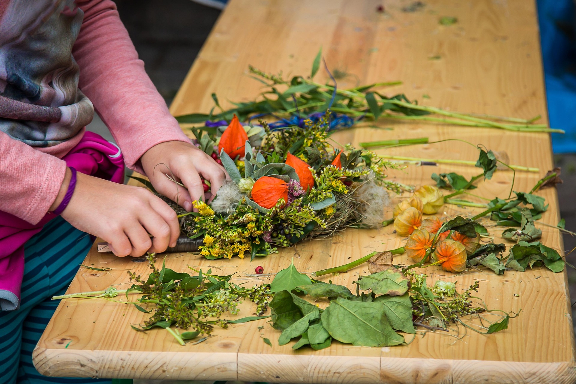 A young child's hands constructs a natural wreath made from fresh greenery and flowers on a wood tabletop.