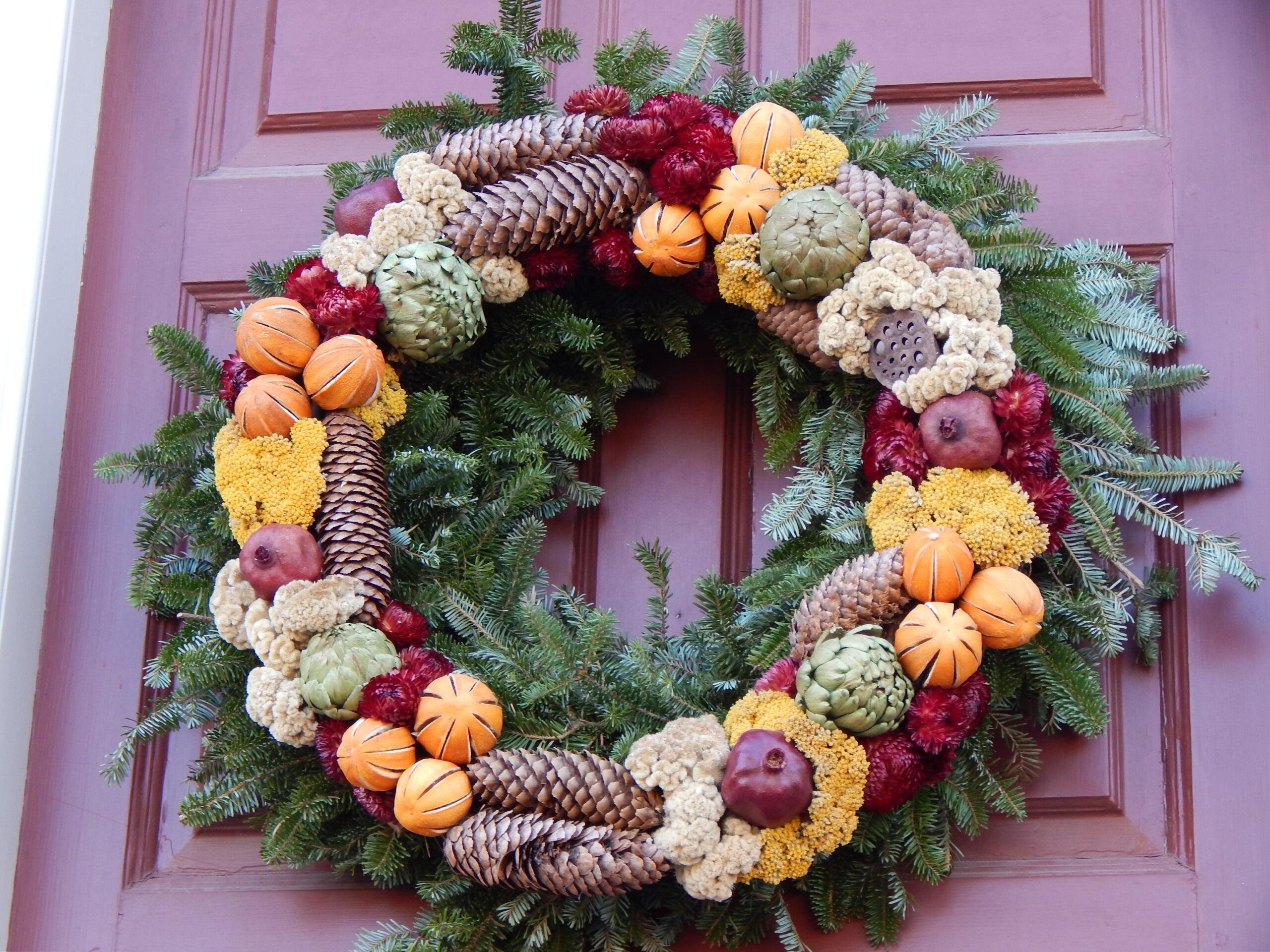 A natural wreath, with green boughs, pinecones, and other natural elements, hangs on a burgundy door.