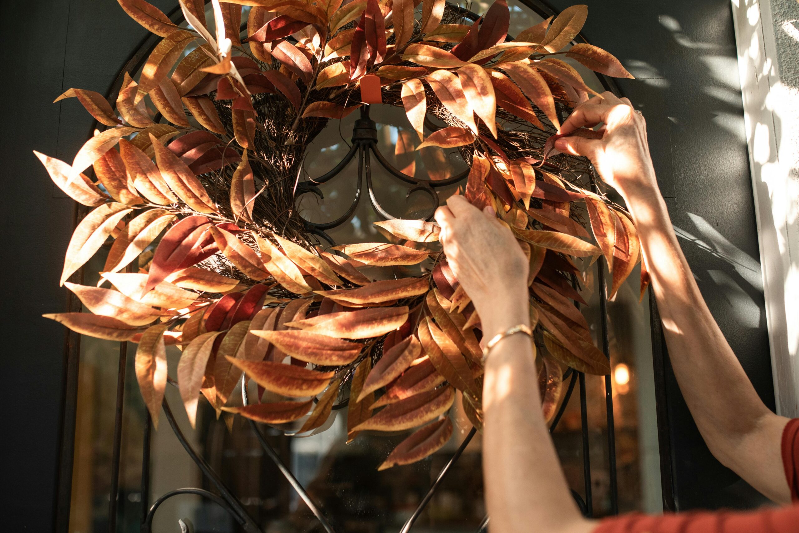 A woman's hands are shown hanging a dried wreath with brown leaf arrangement on a door.