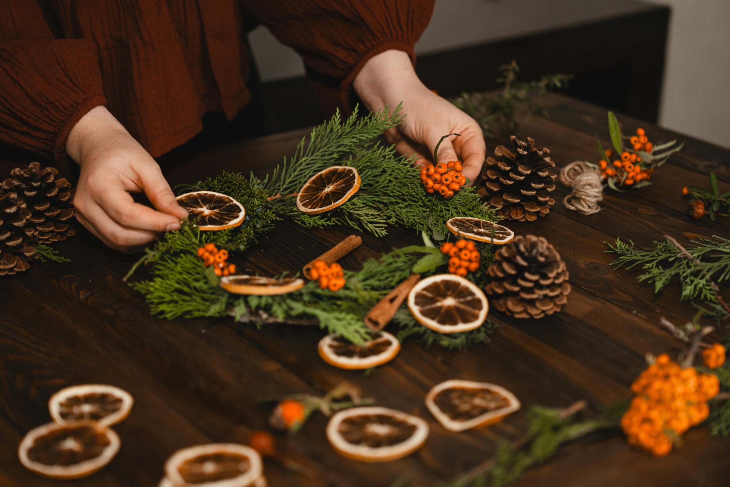 A woman places dried berries and orange slices on a natural bough wreath, constructing it on a wood tabletop.