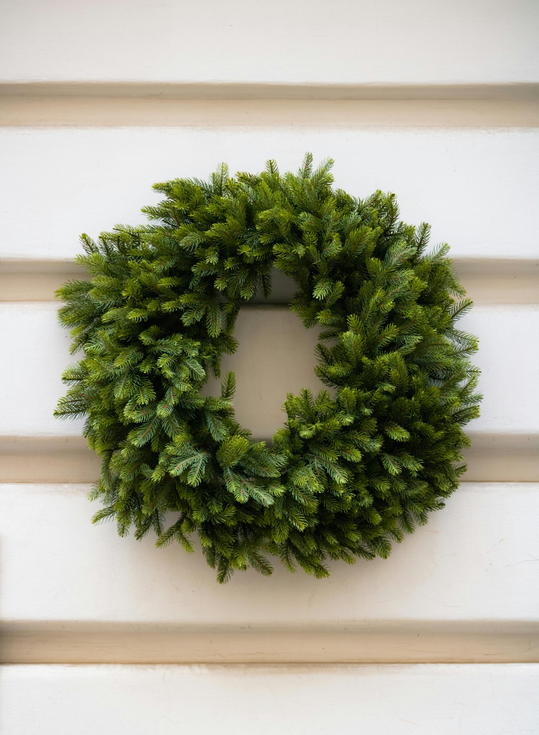 A full, lushly green natural wreath hangs on the white exterior siding of a house.