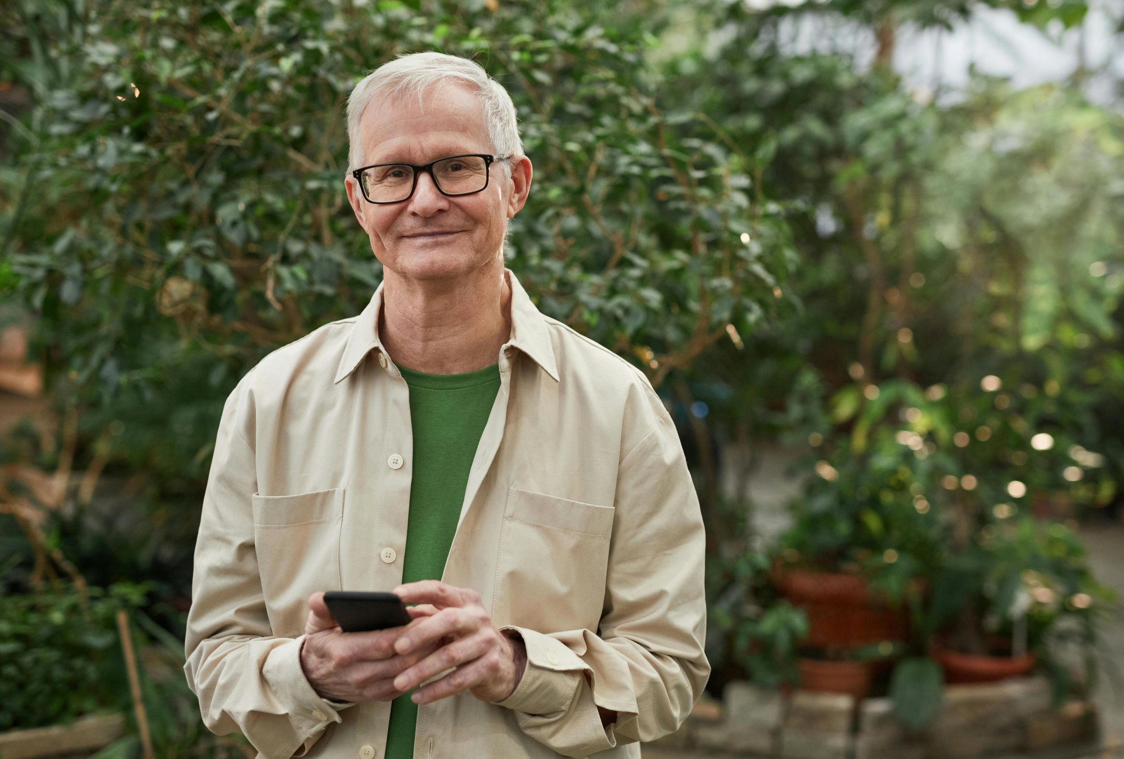 An older man looks contentedly at the camera, smiling, as he holds his phone, with greenery in the background.