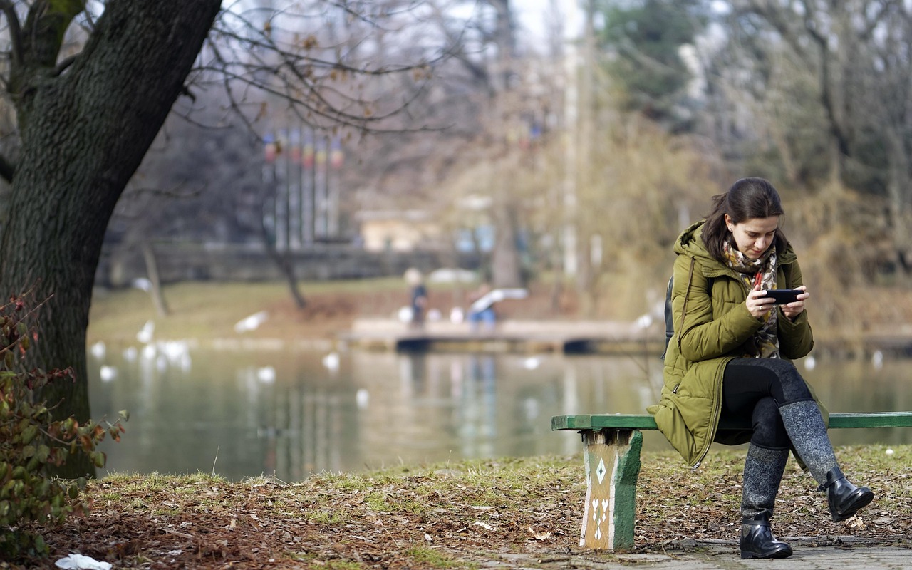 A woman sits on a bench, looking down at her phone, in a park-like setting. 