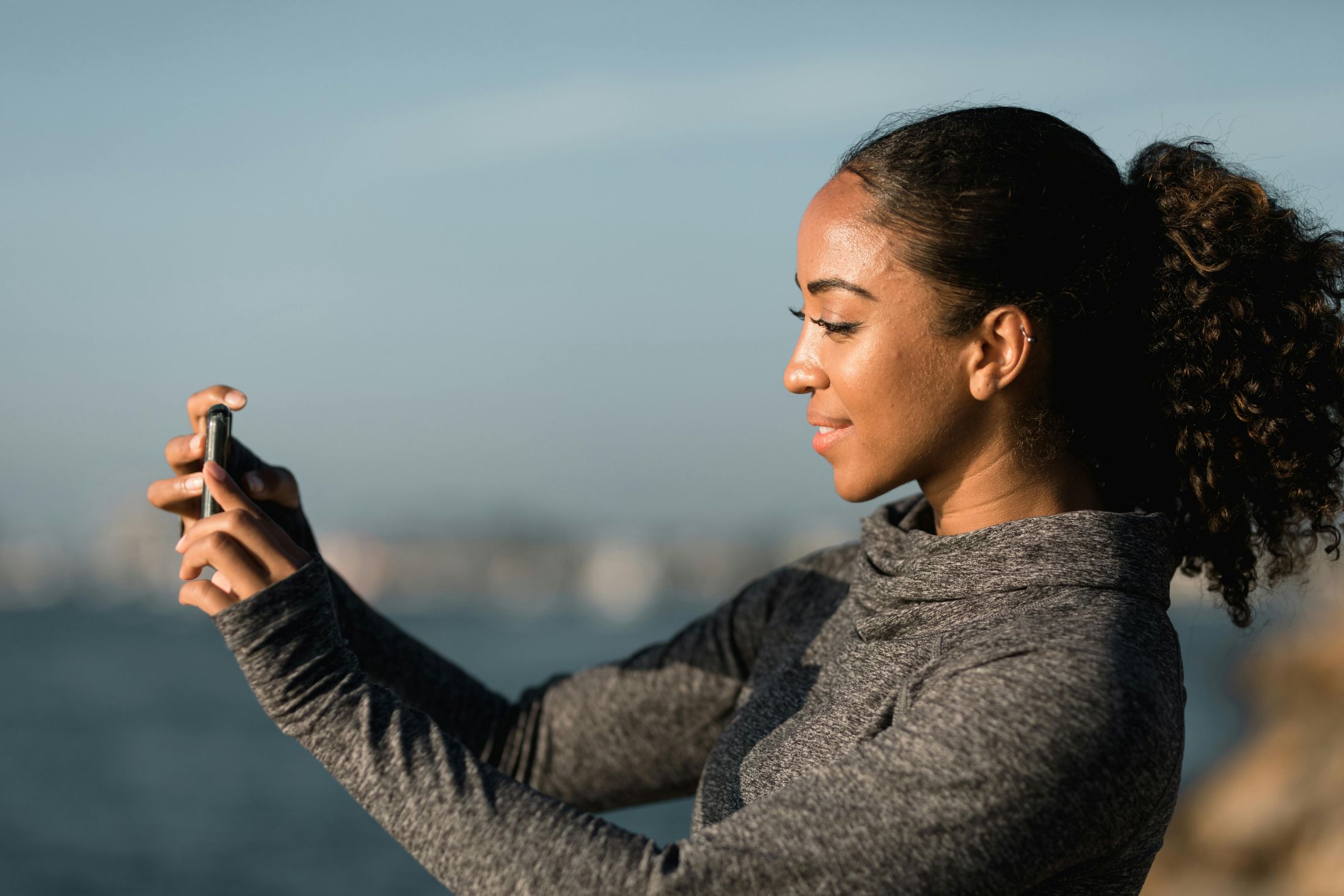 A young Black woman smiles as she takes a photo with her smartphone, a large body of water in the fuzzy background.