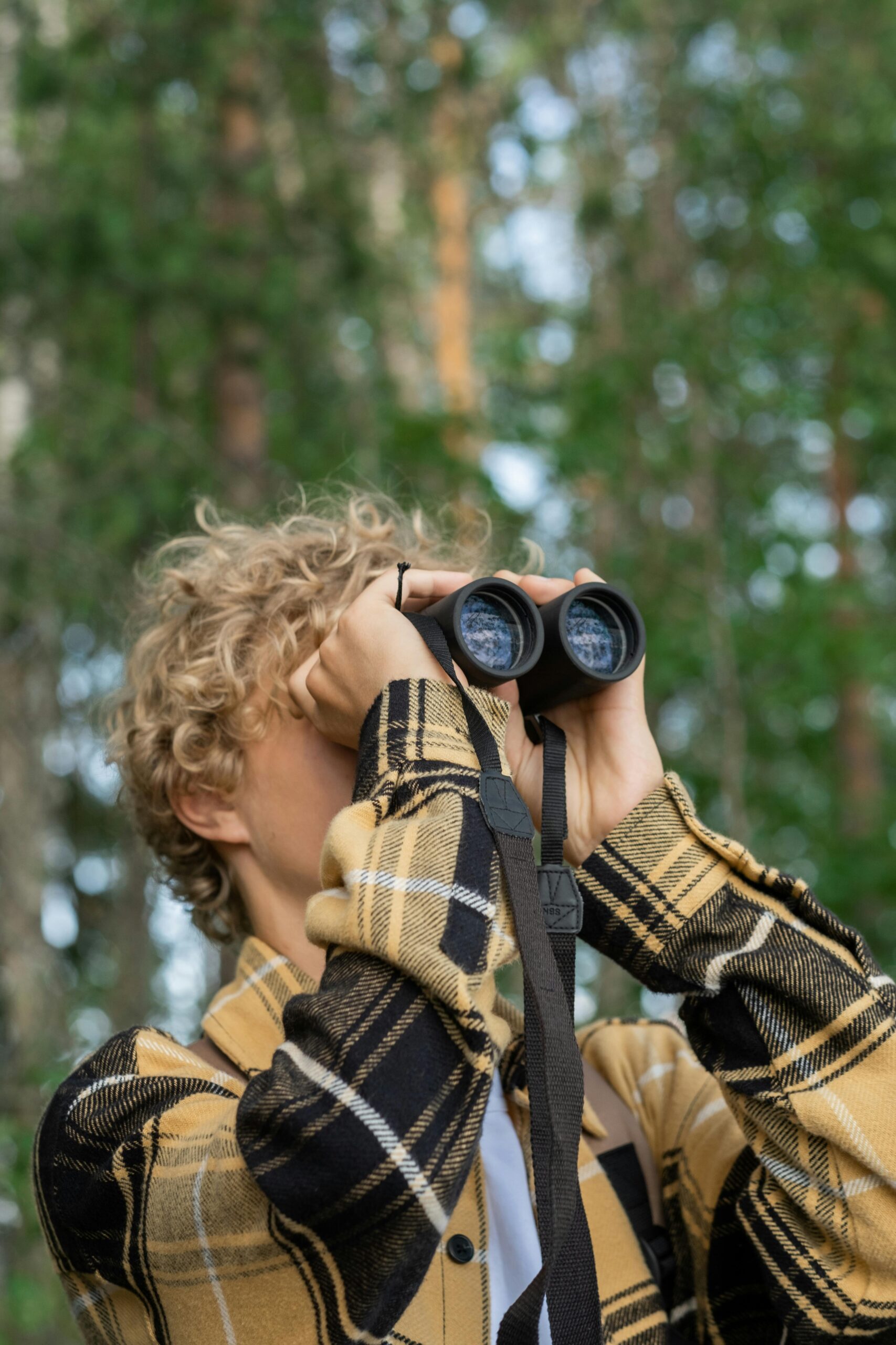 A blonde, curly-haired teenage boy with a yellow and black flannel shirt looks up through binoculars at a tree in a forest setting.