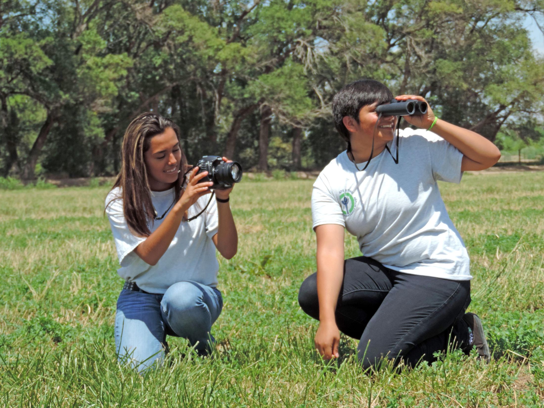 Two teenage girls kneel in a grassy field, forest around them, as they smile and look through binoculars.