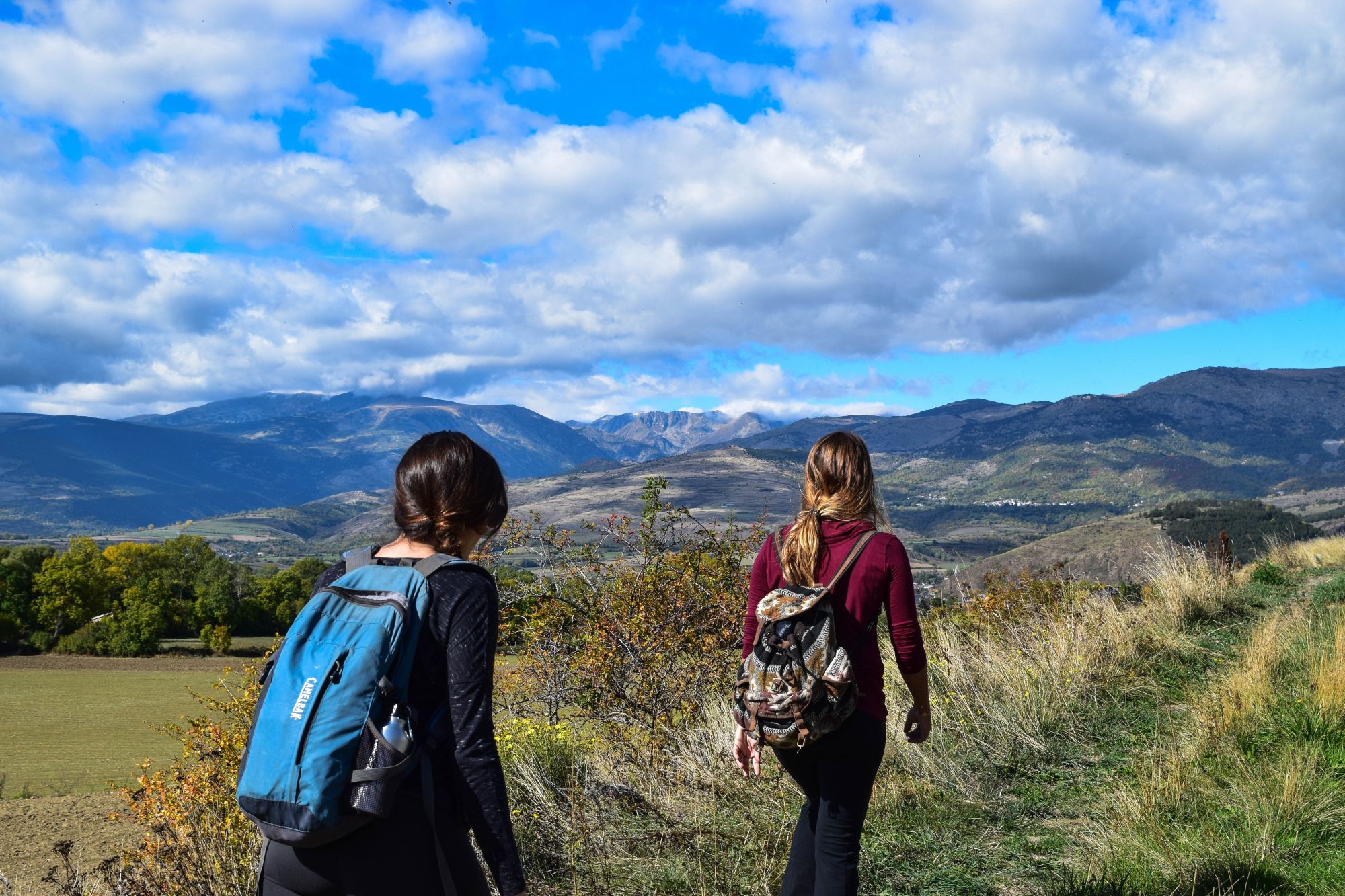 Two teenage girls walk through a natural landscape, mountains and blue sky in background.